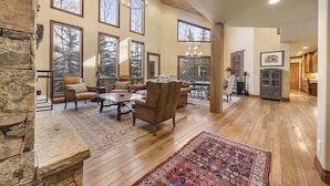 A spacious living room with a large wooden table, leather chairs, and a red patterned rug. There are multiple large windows offering views of trees.