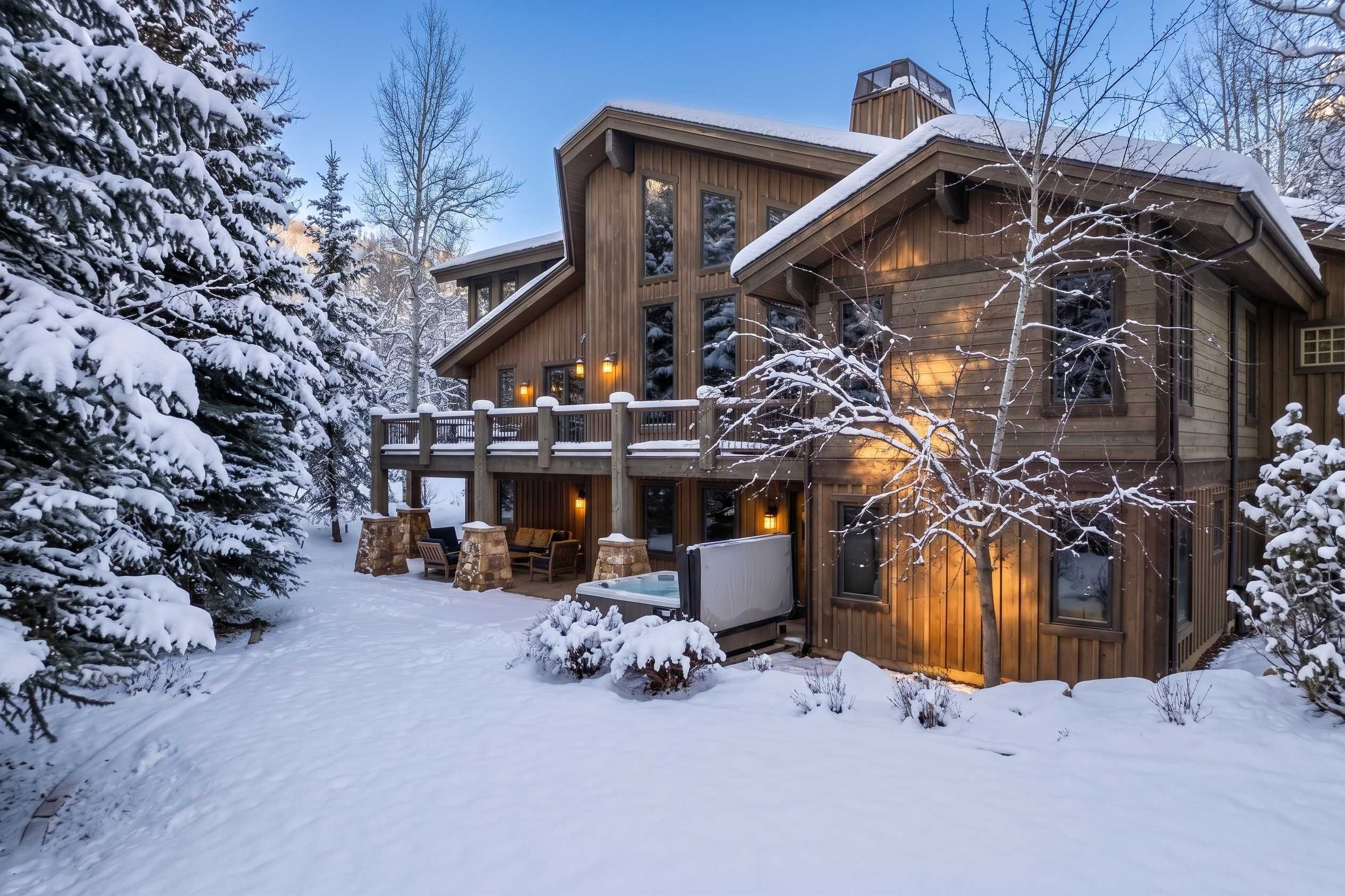Una cabaña grande de madera con un porche cubierto y un jacuzzi, rodeada de árboles cubiertos de nieve.