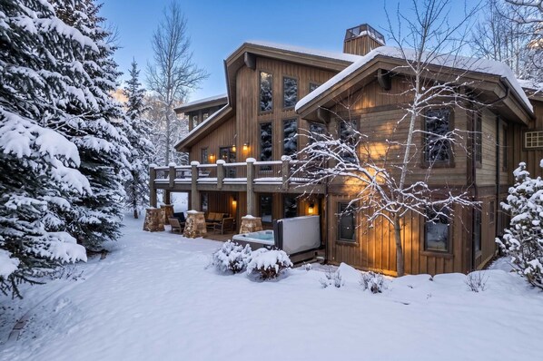 A large wooden cabin with a covered patio and hot tub, surrounded by snow-covered trees.