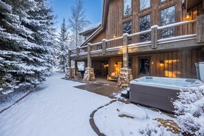A large wooden lodge with a covered patio, stone pillars, and a hot tub.