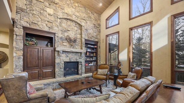 A spacious living room with a stone fireplace, leather furniture, and large windows offering a view of trees.