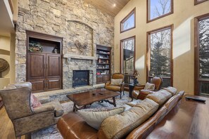 A spacious living room with a stone fireplace, leather furniture, and large windows offering a view of trees.