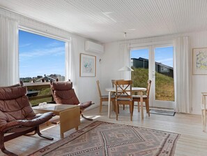 A spacious room with a large window, a dining table with chairs, a brown leather sofa, and a patterned rug. - 5 Star Holiday Home in Lokken (Lokken)