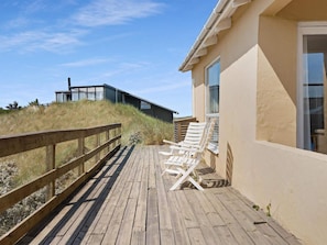 A wooden deck with lounge chairs, a building with large windows, and a grassy hill in the background. - 5 Star Holiday Home in Lokken (Lokken)