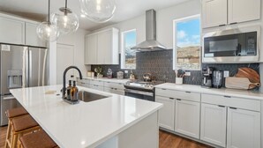 A modern kitchen with white cabinetry, a central island, stainless steel appliances, and a large window providing a view of the outdoors. - The Foxtrot at Mesa Arch Lane (Mountain Green)