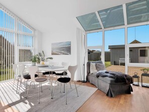 A spacious living area with a dining table, chairs, and a sofa. There is a large window providing a view of the outdoors. - 5 Star Holiday Home in Lokken (Lokken)