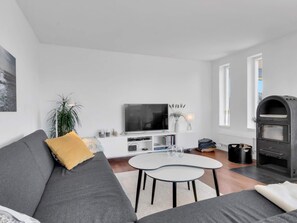 A modern living room with a grey sofa, a small white coffee table, a flat-screen TV, a fireplace, and a large window.