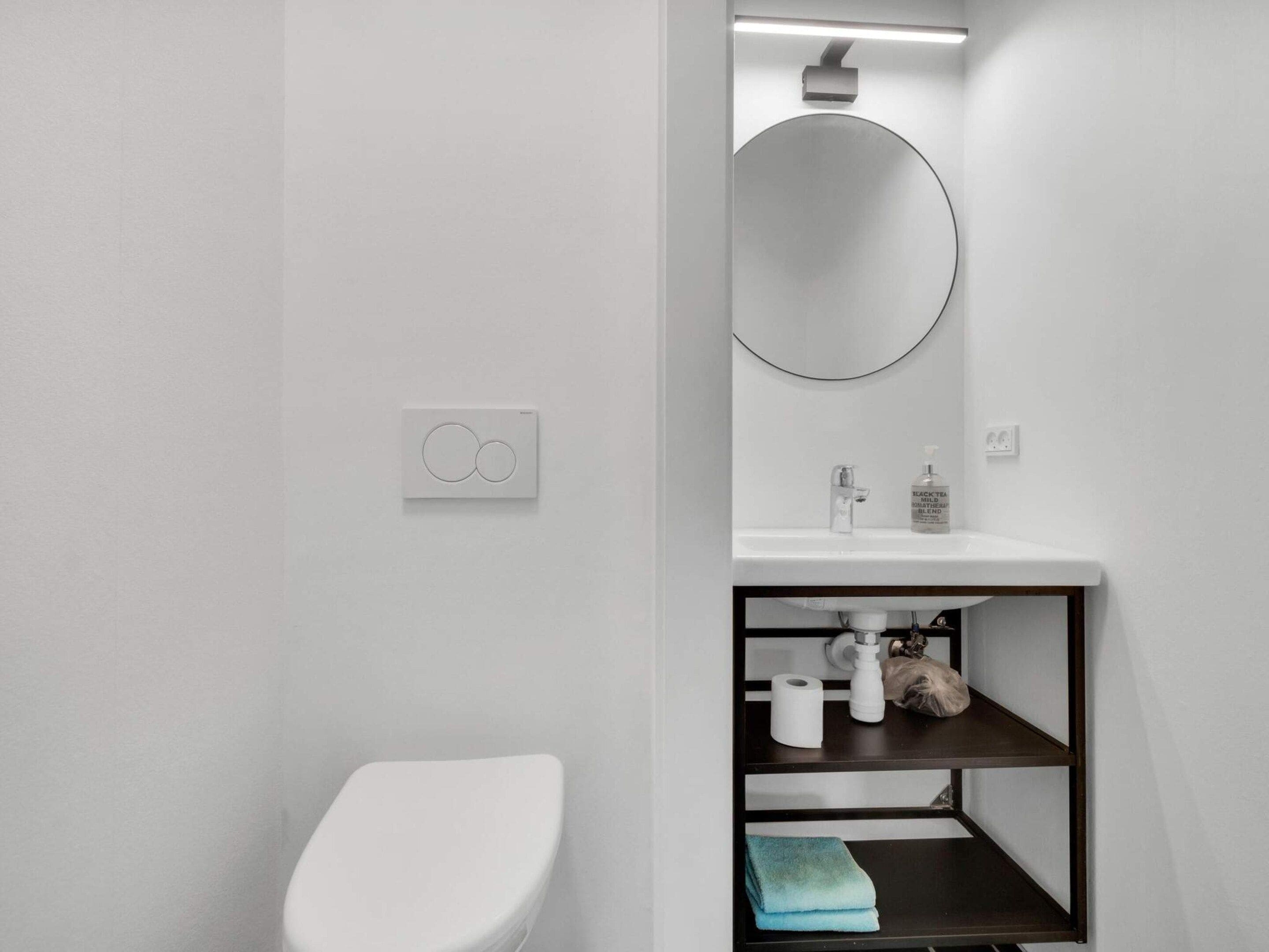 Un baño moderno con inodoro, lavabo, espejo y repisa con artículos de aseo.