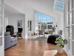 A spacious living room with a fireplace, a dining area with a table and chairs, and a large window providing natural light. - 5 Star Holiday Home in Lokken (Lokken)