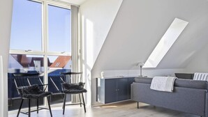 A modern loft bedroom with a sloped ceiling, large windows, a grey sofa, two black chairs, and a wooden floor. - 5 Star Holiday Home in Lokken (Lokken)