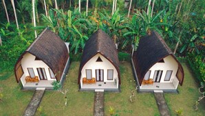 Três casas no estilo de bangalô, com telhados de palha, dispostas em linha com um caminho central. Cada casa possui uma varanda com assentos e uma pequena mesa.