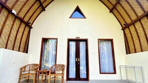 Um quarto com teto alto e abobadado, uma porta de madeira com uma placa acima e duas janelas com cortinas. Há duas cadeiras de madeira e uma mesa pequena no quarto.