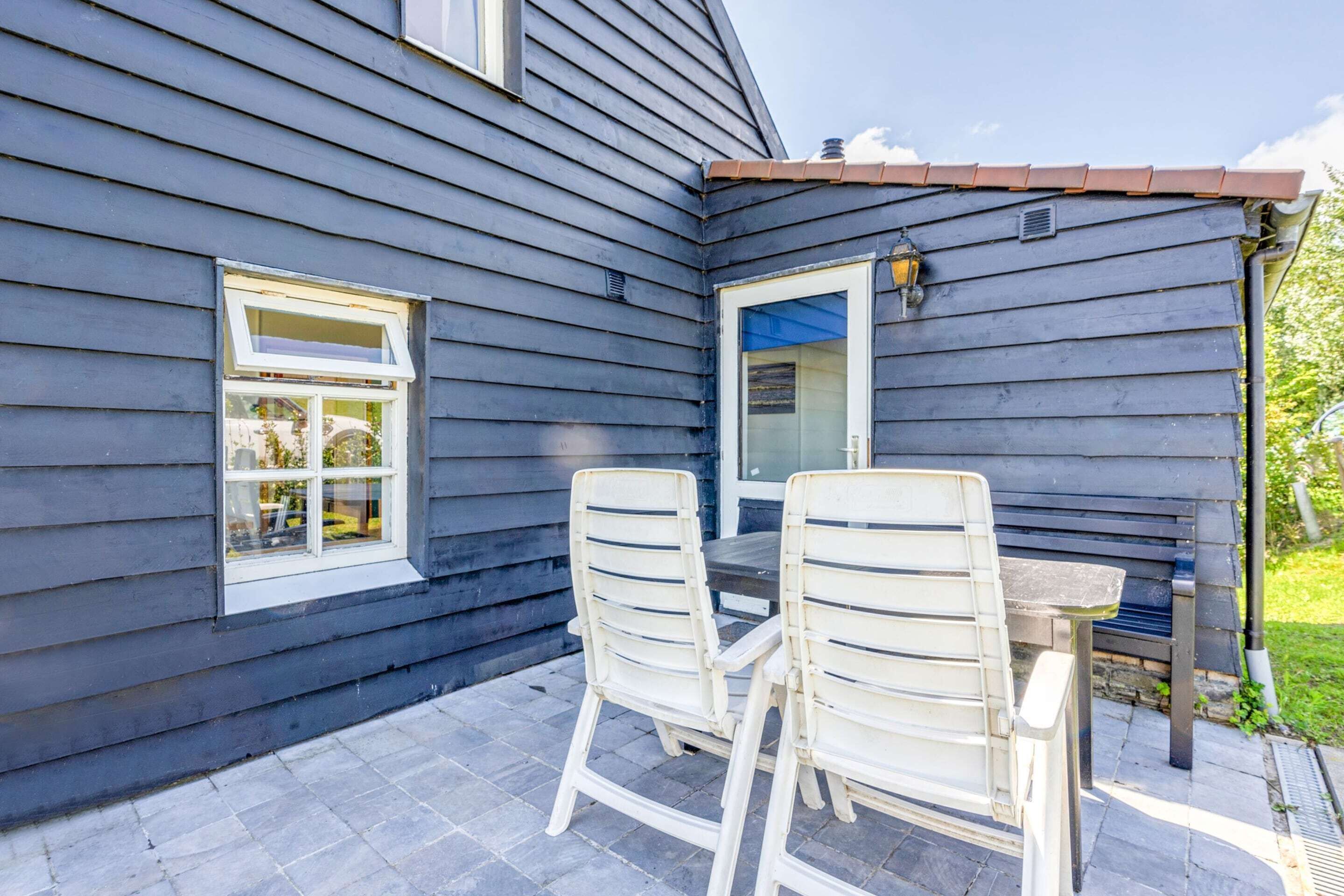 Une terrasse avec deux chaises blanches et une table, une fenêtre et une porte surmontée d’un auvent bleu.