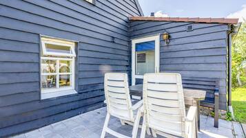 Une terrasse avec deux chaises blanches et une table, une fenêtre et une porte surmontée d’un auvent bleu.