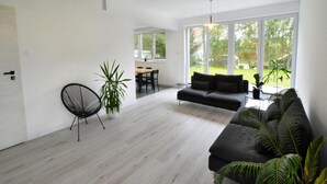 Ein modernes Wohnzimmer mit einer schwarzen Couch, einem Essbereich mit Tisch und Stühlen und großen Fenstern mit Blick ins Freie.