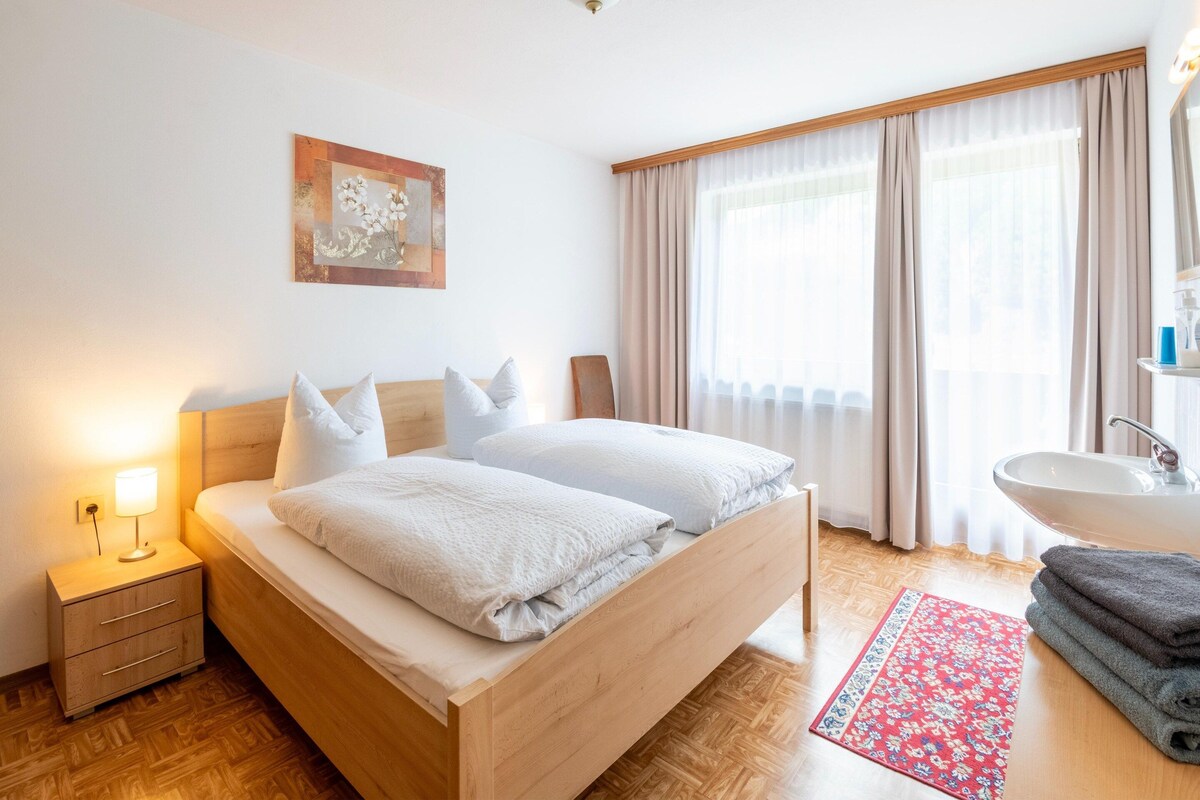 Habitación de hotel con cama de madera, mesita de noche con lámpara, baño con lavamanos y ventana con cortinas.
