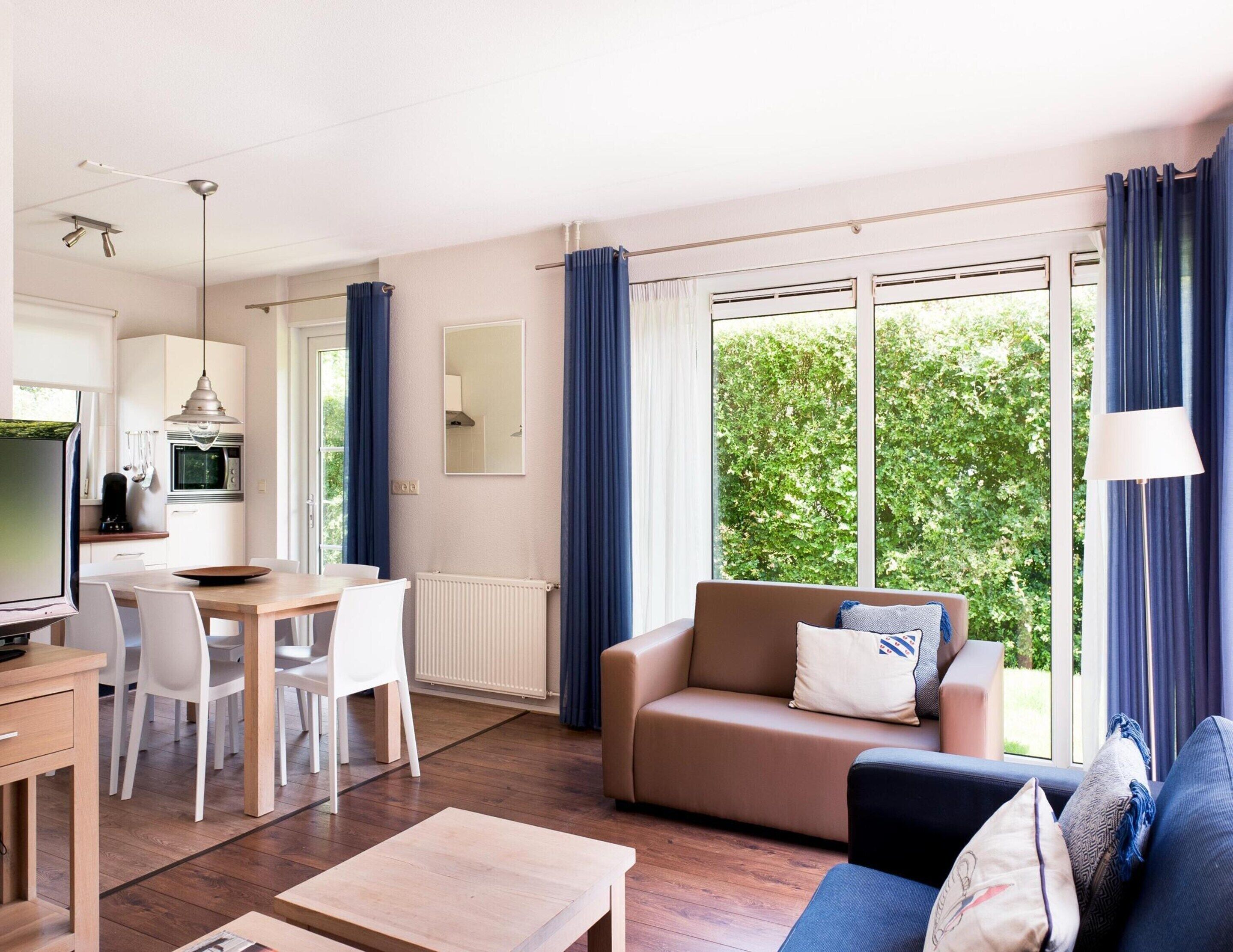Una sala moderna con sofá, mesa de centro y comedor. Hay una cocina con microondas y televisión. Ventanales con cortinas ofrecen vistas a la vegetación exterior.