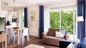 Ein modernes Wohnzimmer mit Sofa, Couchtisch und Essbereich. Es gibt eine Küche mit Mikrowelle und Fernseher. Große Fenster mit Vorhängen bieten Blick auf Grünflächen.