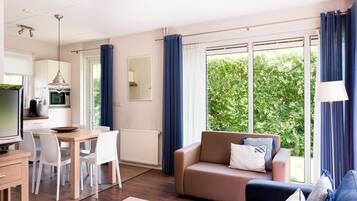Una sala moderna con sofá, mesa de centro y comedor. Hay una cocina con microondas y televisión. Ventanales con cortinas ofrecen vistas a la vegetación exterior.