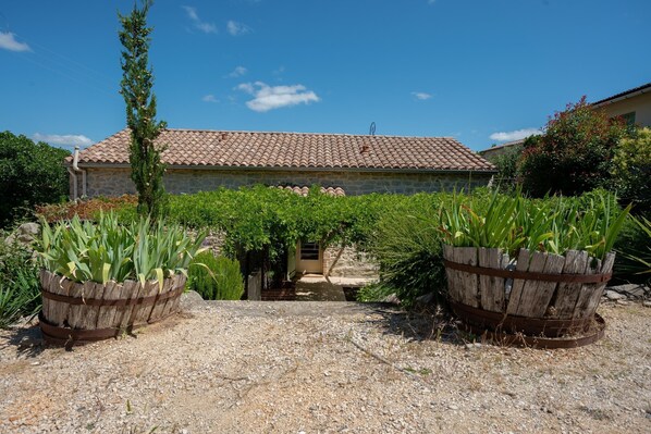 A stone building with a tiled roof, a gravel path leading to a door, and two wooden planters with green plants.