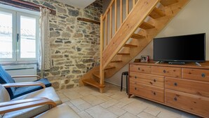 A room with a stone wall, wooden staircase, a flat-screen TV, a wooden dresser, and a window with curtains. - Pretty Holiday Home in Courry With Swimming Pool (Courry)
