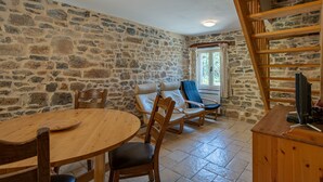 A stone-walled room with a round wooden table, two wooden chairs, a blue cushioned chair, a wooden TV stand, and a staircase. - Pretty Holiday Home in Courry With Swimming Pool (Courry)