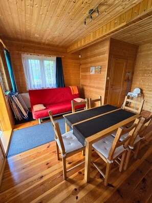 A wooden cabin interior with a dining table, chairs, a red sofa, and a window with curtains.