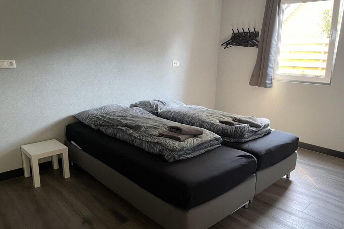 Un lit simple avec un matelas noir et une literie grise, une petite table de chevet blanche, un porte-manteau fixé au mur et une fenêtre avec des rideaux.