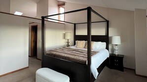 Una cama con dosel acompañada de un banco a juego, una mesita de noche con lámpara y una puerta que da a otra habitación.