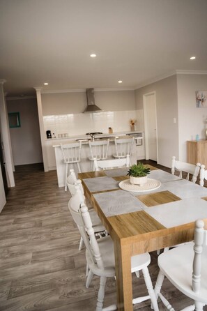 Een moderne eetruimte met een houten tafel en witte stoelen, een keuken op de achtergrond en een houten vloer.