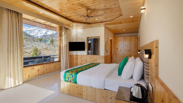 A wooden-paneled bedroom with a bed, a window with curtains, a mounted TV, and a view of mountains. - Treebo Premium Silverstone Resort With Mountain Valley View (Manali)