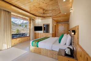 Une chambre à coucher aux murs lambrissés, avec un lit, une fenêtre avec des rideaux, un téléviseur fixé au mur et une vue sur les montagnes.