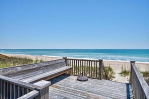 Terraza de madera con un banco y una mesa, con vista a una playa y olas.
