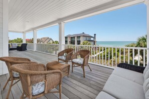Une terrasse extérieure couverte, aménagée avec des meubles en osier, une balustrade blanche et offrant une vue sur l’océan et une maison.