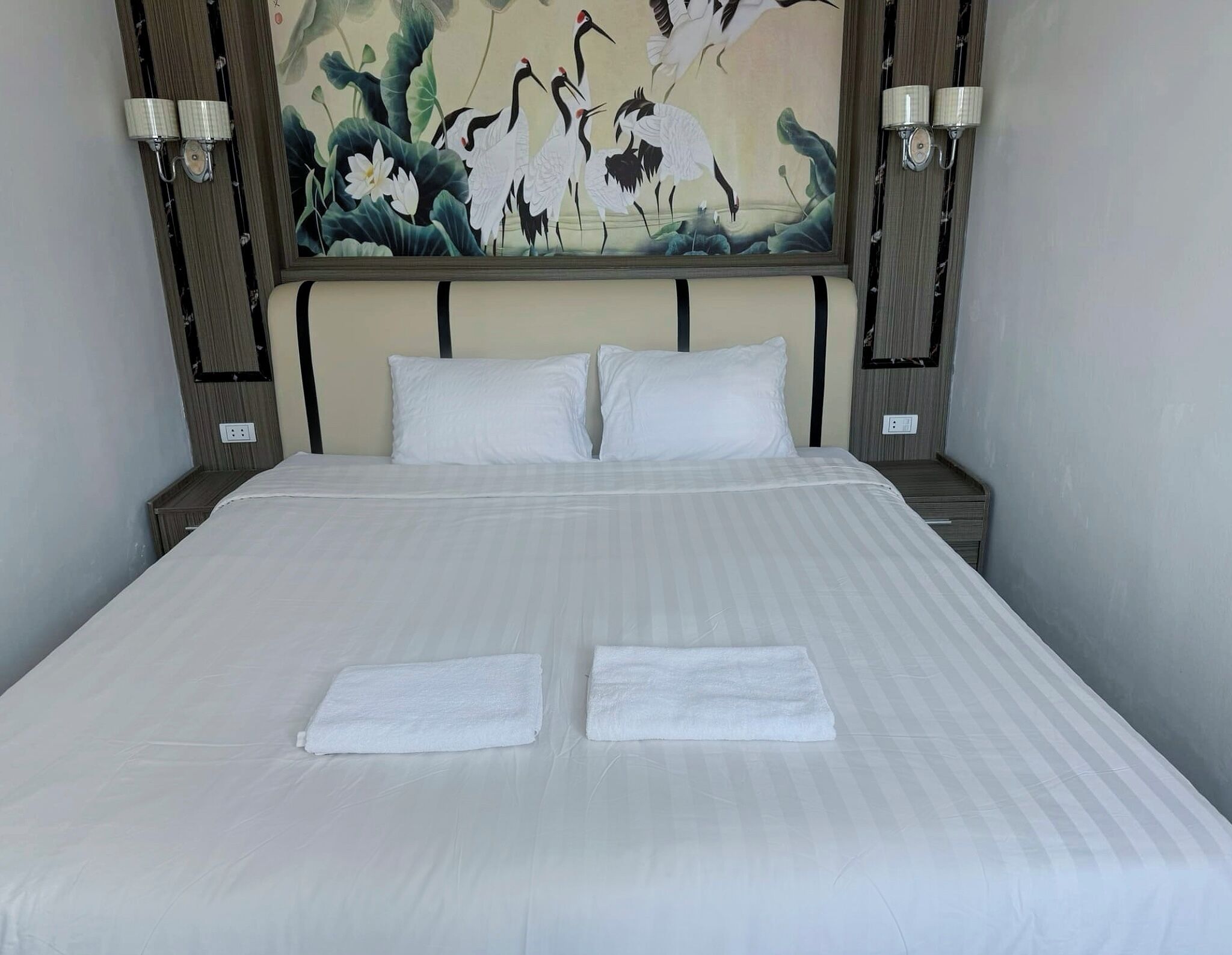 Un lit bien fait, avec du linge de lit blanc et deux serviettes, une grande peinture murale représentant des oiseaux et du feuillage, et deux lampes fixées au mur.