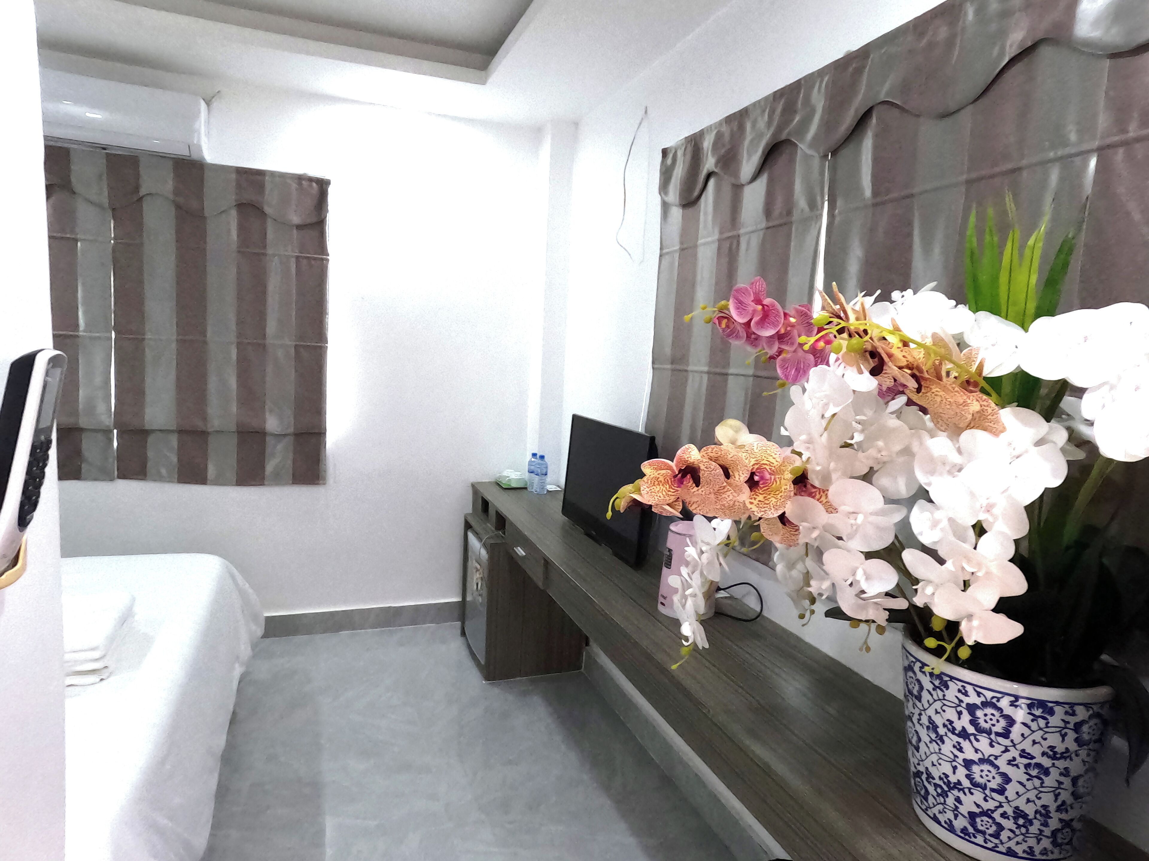 A hotel room with a bed, a TV on a wooden stand, a potted orchid plant, and a window with striped curtains.