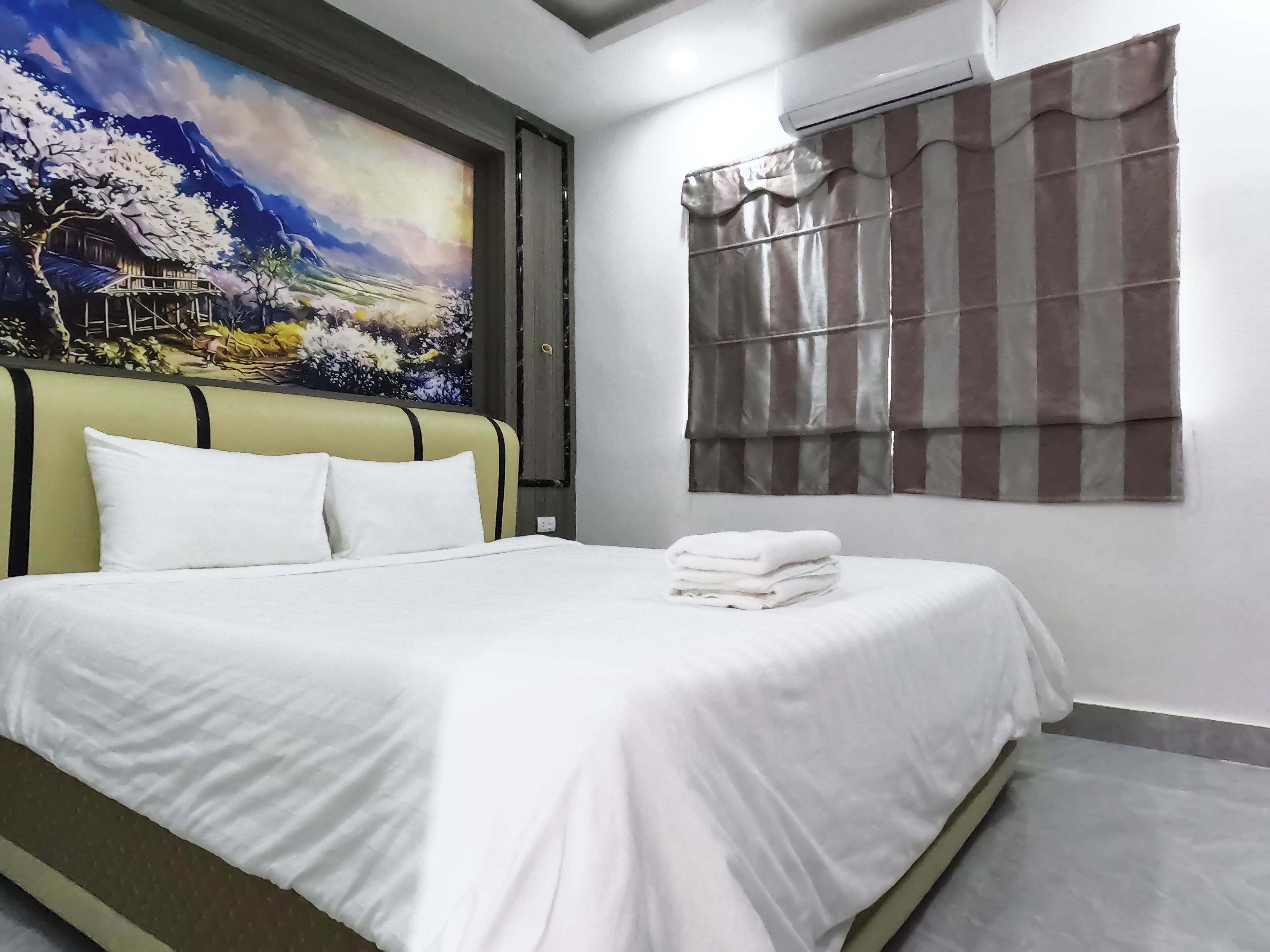A neatly made bed with white linens and pillows, a large painting of a mountainous landscape on the wall, and a window with striped curtains.