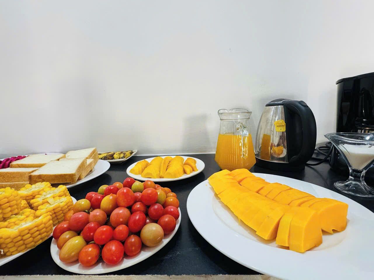 A breakfast buffet with plates of corn, tomatoes, mango slices, and juice.