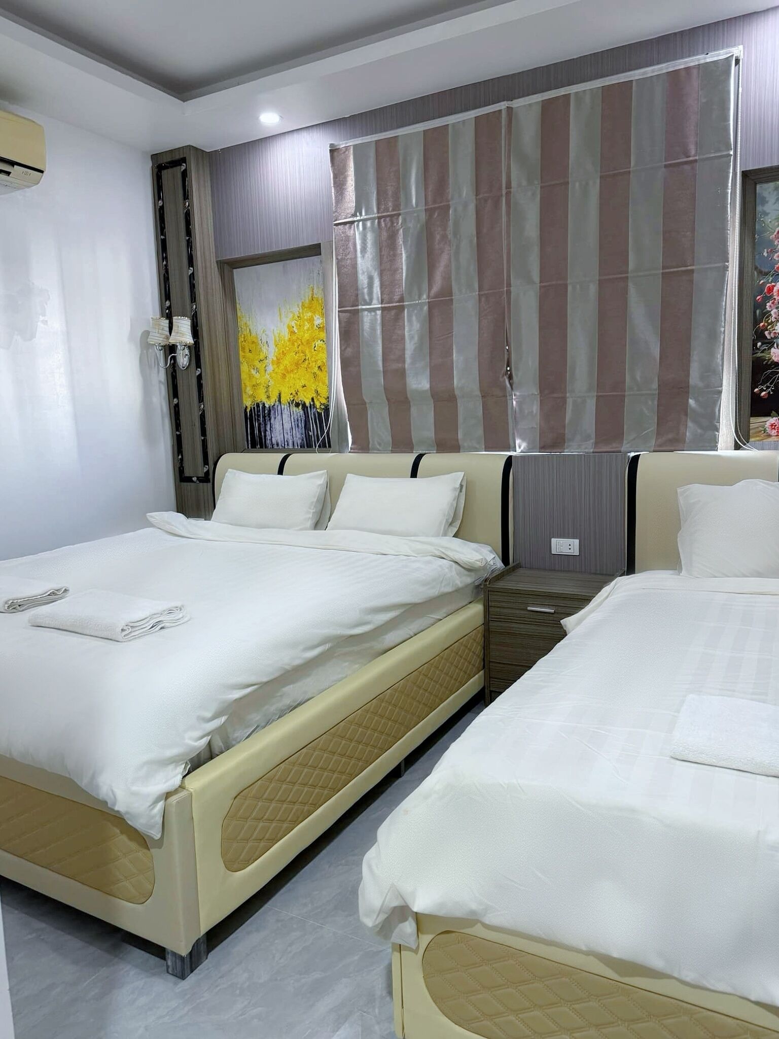 Habitación de hotel con dos camas, un ventanal con cortinas a rayas, un cuadro de flores amarillas y un aire acondicionado de pared.
