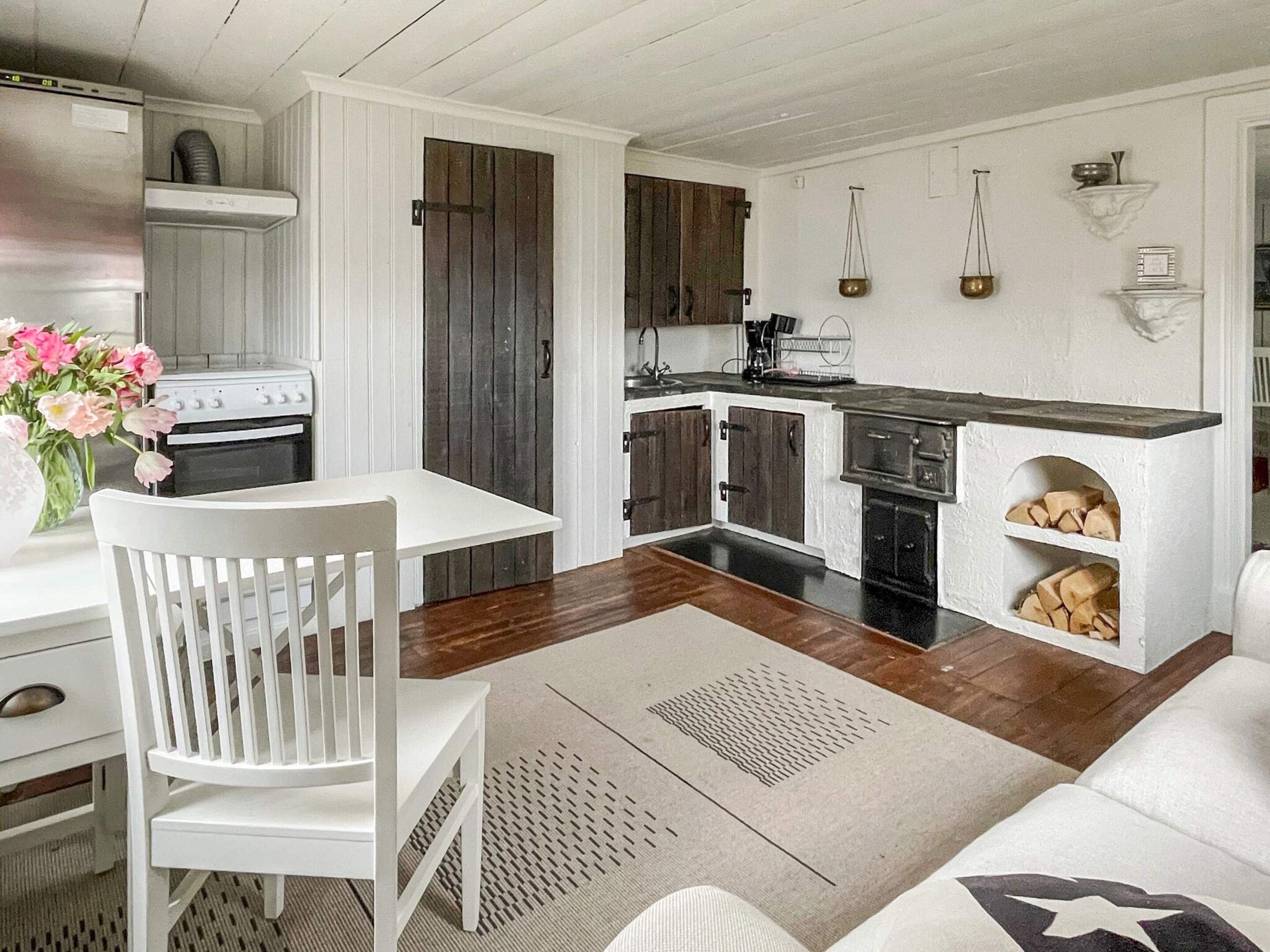 Una cocina compacta con muebles blancos, una mesa de comedor con una silla blanca, y un mueble de almacenamiento de madera empotrado.