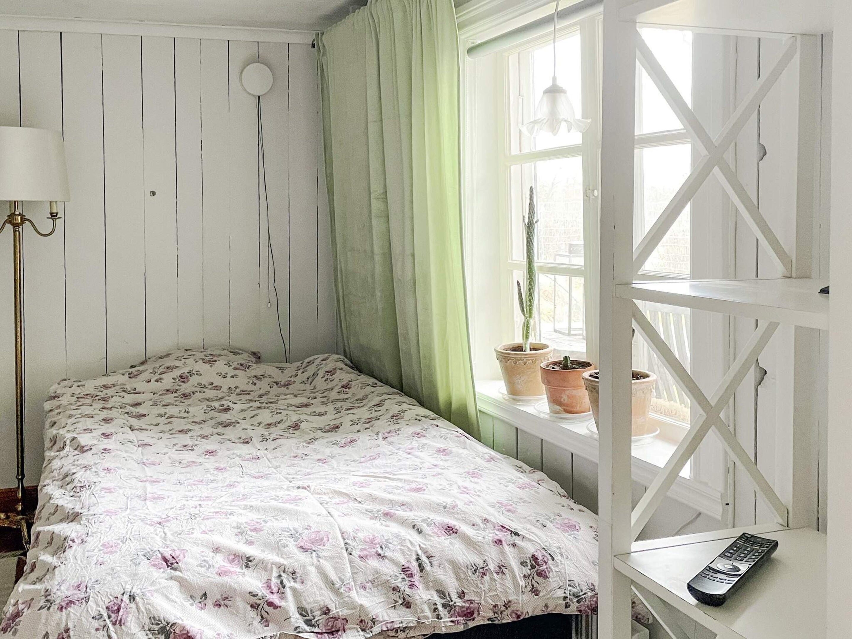 Quarto com cama, janela com cortinas, planta em vaso, luminária e controle remoto em uma prateleira.