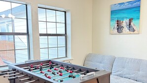 A foosball table in a room with large windows, a gray sofa, and a painting of a beach scene. - Waterfront 3BR 2BA Home near Galveston, Kemah, NASA, NRG, Rodeo, Houston (La Porte)