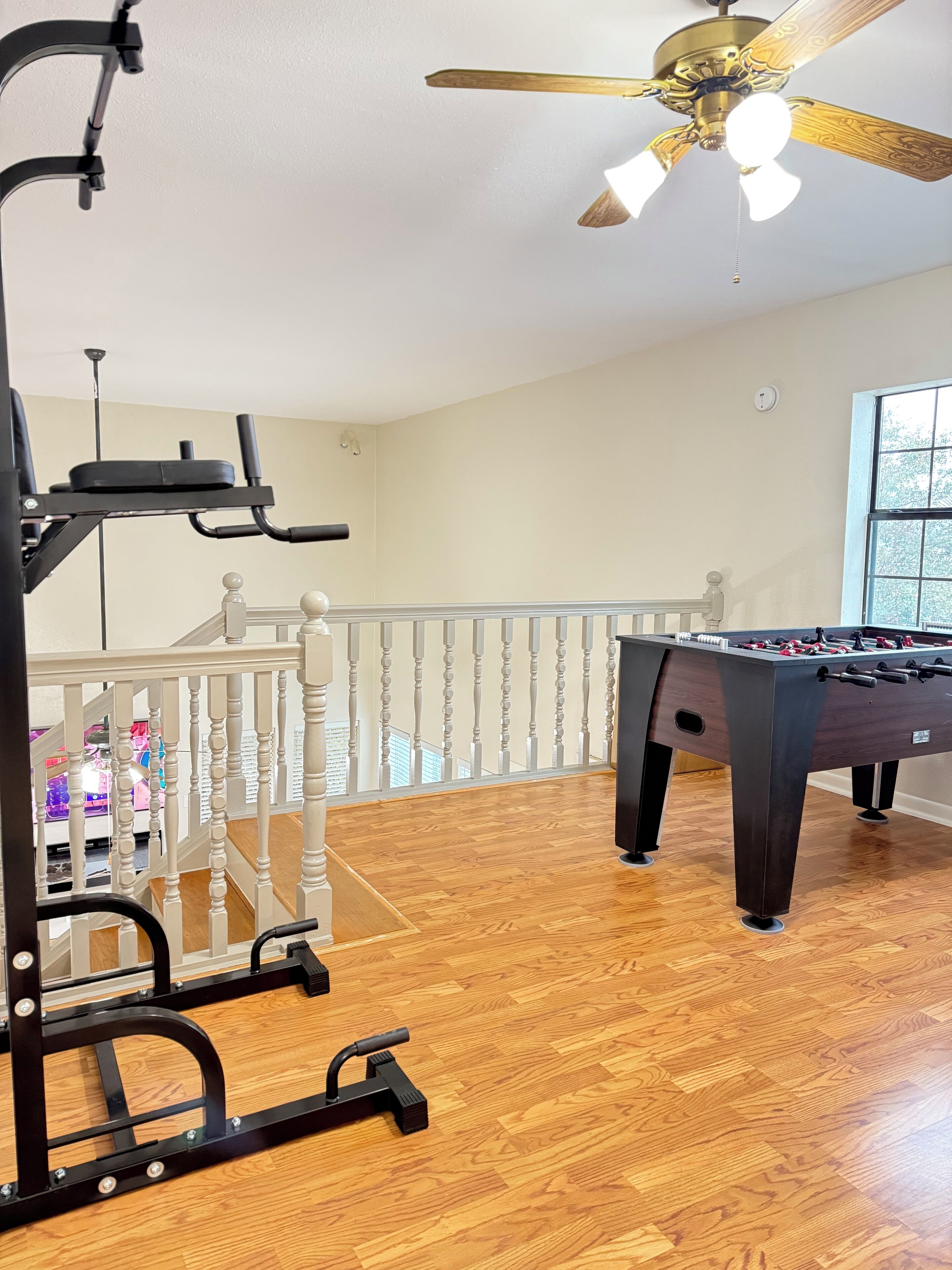 A room with a foosball table, exercise equipment, a ceiling fan, and a balcony with a railing.