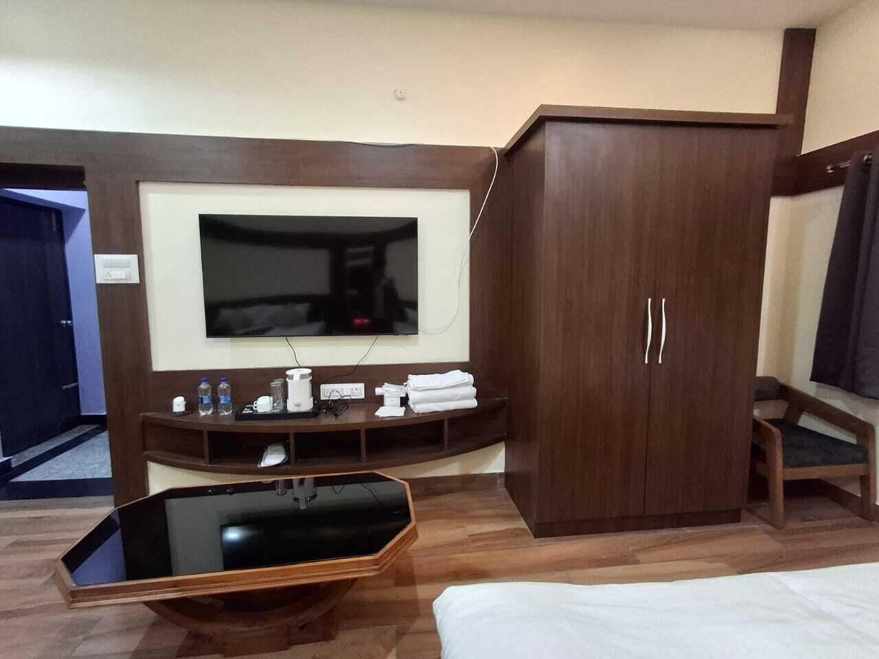 A hotel room with a large flat-screen TV mounted on the wall, a wooden wardrobe, a glass-top coffee table, and a bed with white bedding.