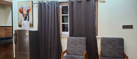 A compact kitchen with a refrigerator, two chairs, and a window with curtains.