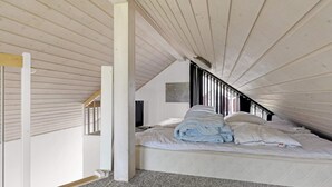 A bedroom with a sloped ceiling, a bed with white bedding, a television on the wall, and a window with blinds. - 5 Star Holiday Home in Lokken (Lokken)