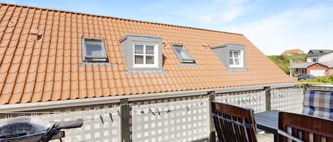 A residential building with a sloped roof, two small windows, and a wooden deck with outdoor furniture.