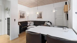 A modern kitchen with a wooden ceiling, a dining area with a white table and dark chairs, and a dark leather sofa with floral cushions. - 5 Star Holiday Home in Lokken (Lokken)
