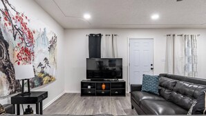 Ein Wohnzimmer mit schwarzem Ledersofa, einem Fernseher auf einem schwarzen Standfuß, einem Gemälde an der Wand und einer Tür mit Vorhängen.