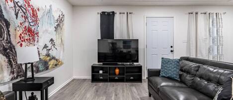 A living room with a black leather sofa, a TV on a black stand, a painting on the wall, and a door with curtains.